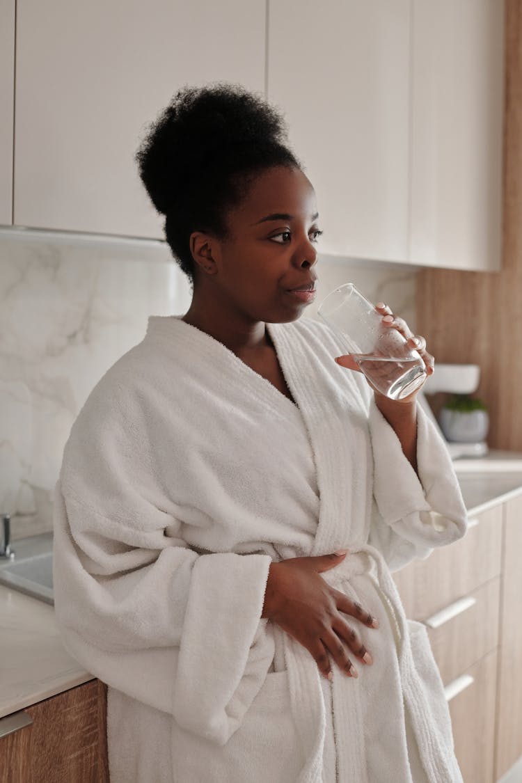 Woman In Bathrobe Standing In The Kitchen Drinking Water