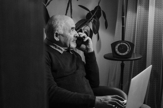 A senior adult man in a cozy indoor setting speaks on the phone while using his laptop.