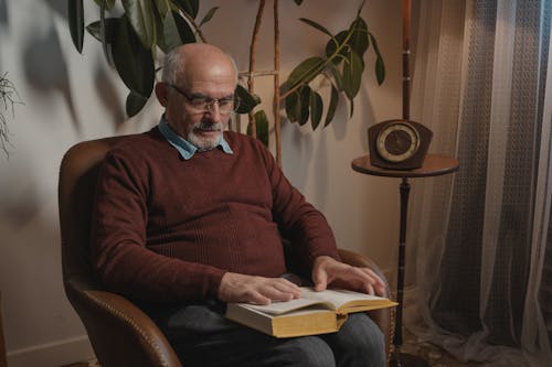 Man in Brown Sweater Wearing Eyeglasses while Reading Book