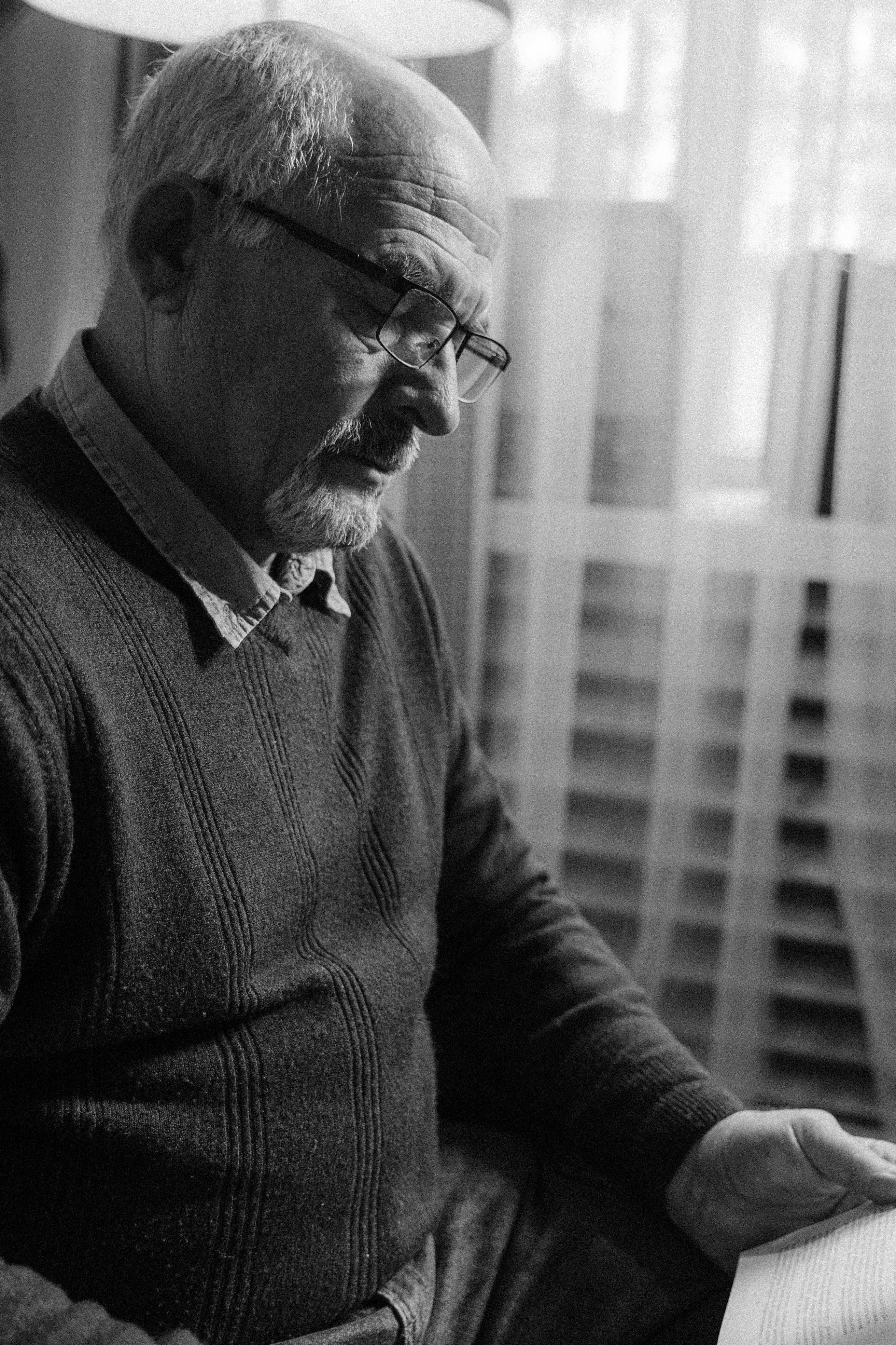 A senior man wearing eyeglasses reads in a cozy indoor setting, captured in black and white.