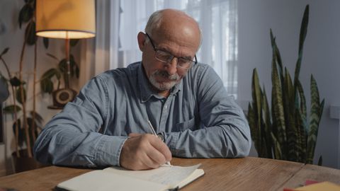 A senior adult man writing in a notebook at a desk in a cozy home office setting.