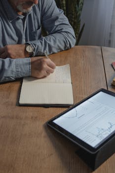 Man writing notes at home desk with tablet showing graphs, illustrating remote work.
