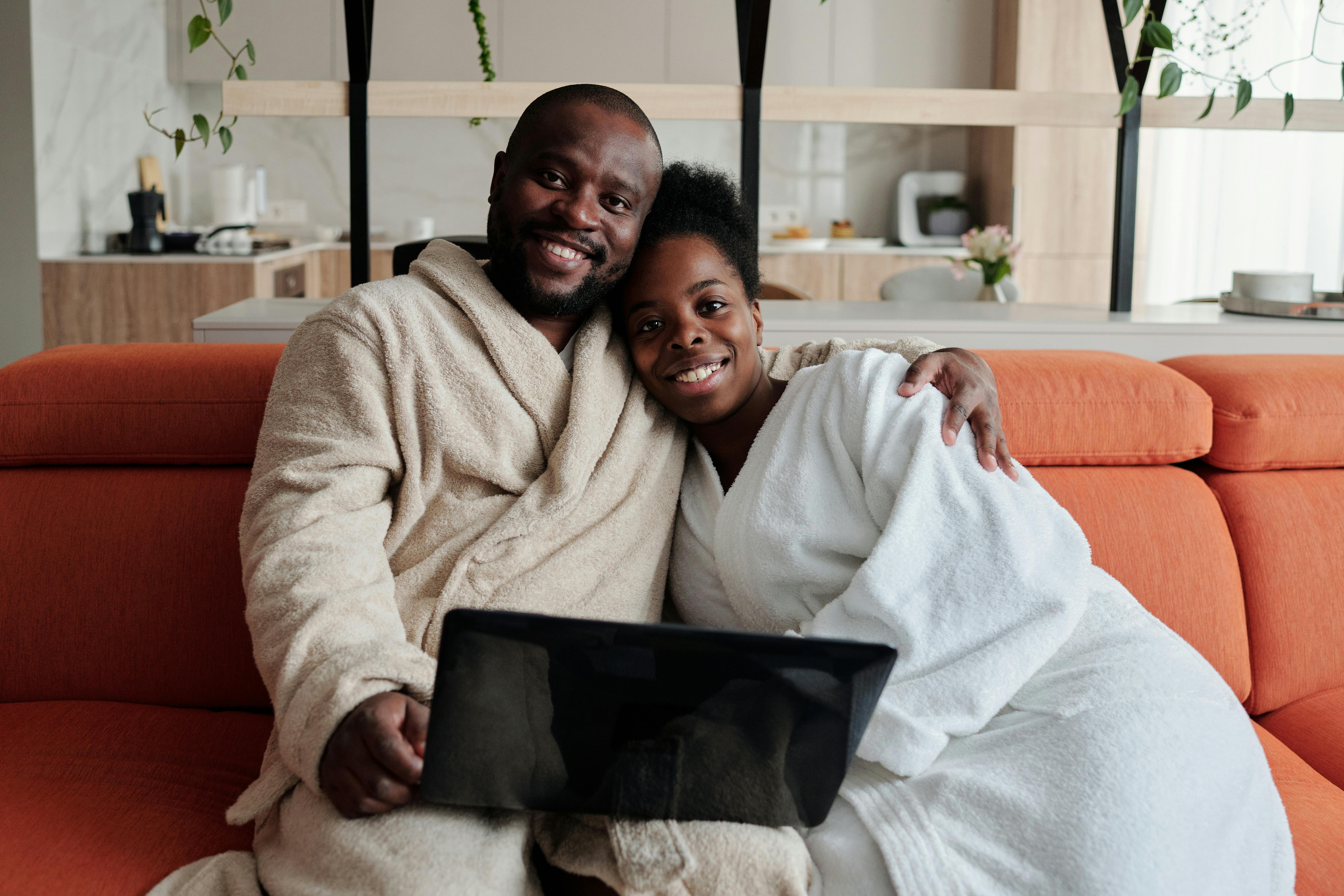 Smiling couple in bathrobes relaxing on a couch with a laptop, embracing warmth and togetherness.