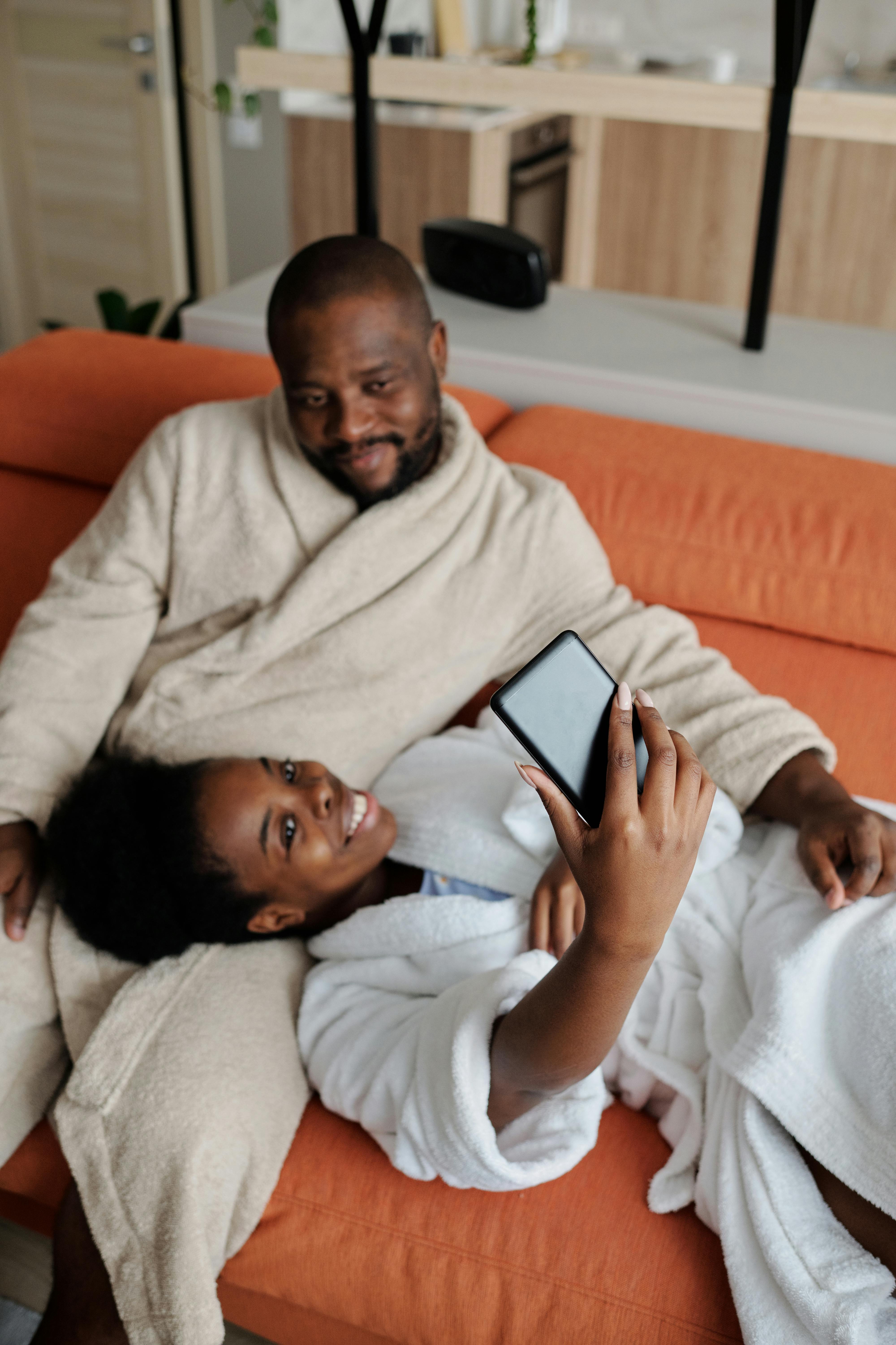 A cheerful couple in bathrobes snapping a selfie on a cozy couch indoors.