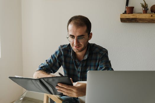 A man with glasses writes in a notebook while working on his laptop in a home office.