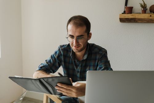 Free A man with glasses writes in a notebook while working on his laptop in a home office. Stock Photo