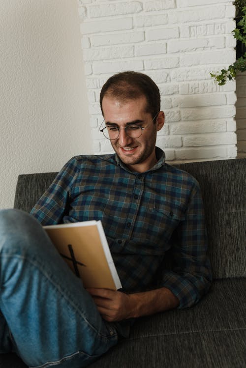 A Man in Plaid Long Sleeves Sitting on Gray Couch Reading a Book 