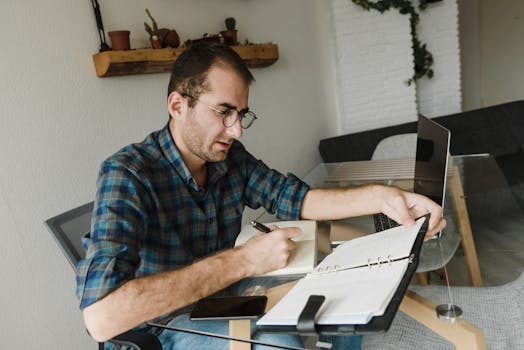 Caucasian male freelancer working from home with laptop, notebook, and pen.