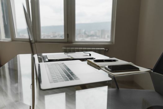 A modern office setup featuring a laptop, notebook, and pen on a glass table with city view.