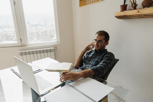 A young man working remotely in his home office, talking on the phone, using a laptop.