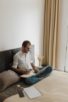Adult man focused on remote work with laptop and notebook on bed. Perfect for productivity themes.