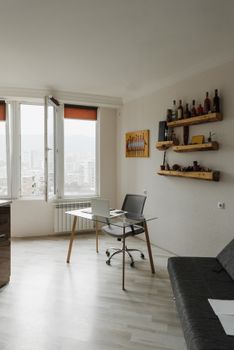 A bright and modern apartment workspace interior with a desk, shelves, and large windows.
