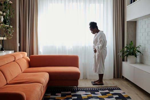 An African American woman in a bathrobe checks her weight on a scale near an orange sofa.