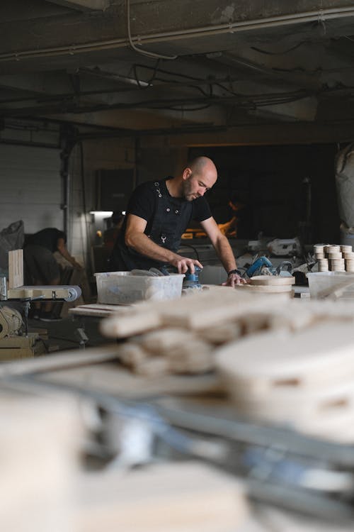 Side view of concentrated male master working with sander at workbench  in professional studio with stack of wooden boards