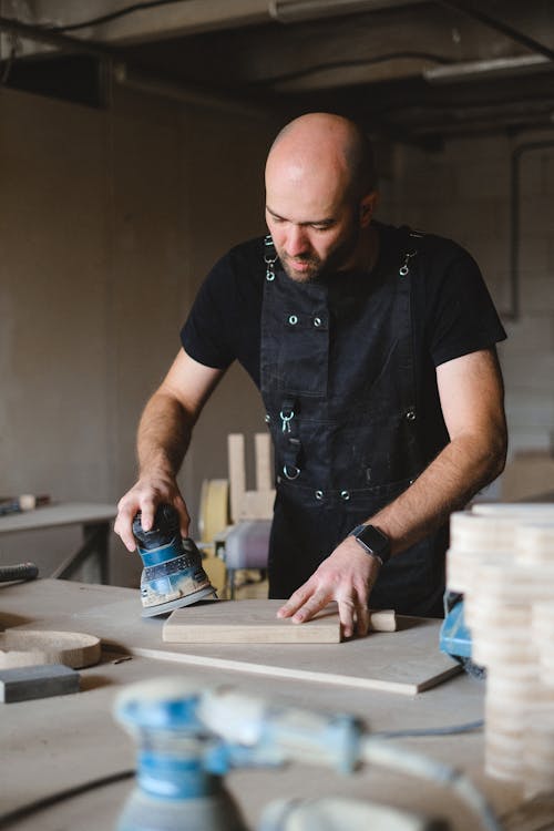 Concentrated bald male woodworker wearing black apron polishing wooden board with special instrument while working at workbench in professional joinery