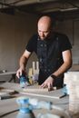 Concentrated bald male woodworker wearing black apron polishing wooden board with special instrument while working at workbench in professional joinery