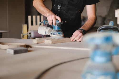 Unrecognizable male woodworker wearing uniform and polishing wooden board with random orbital sander at big table in professional studio