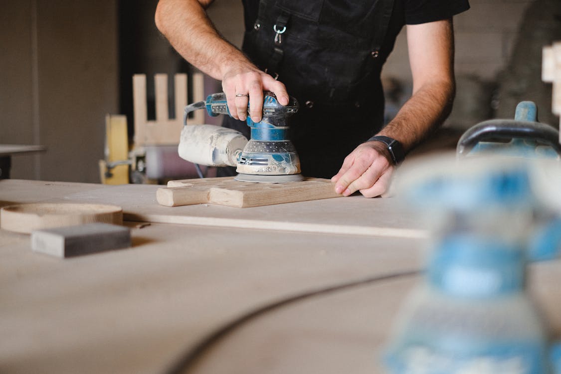 Free Unrecognizable male woodworker wearing uniform and polishing wooden board with random orbital sander at big table in professional studio Stock Photo