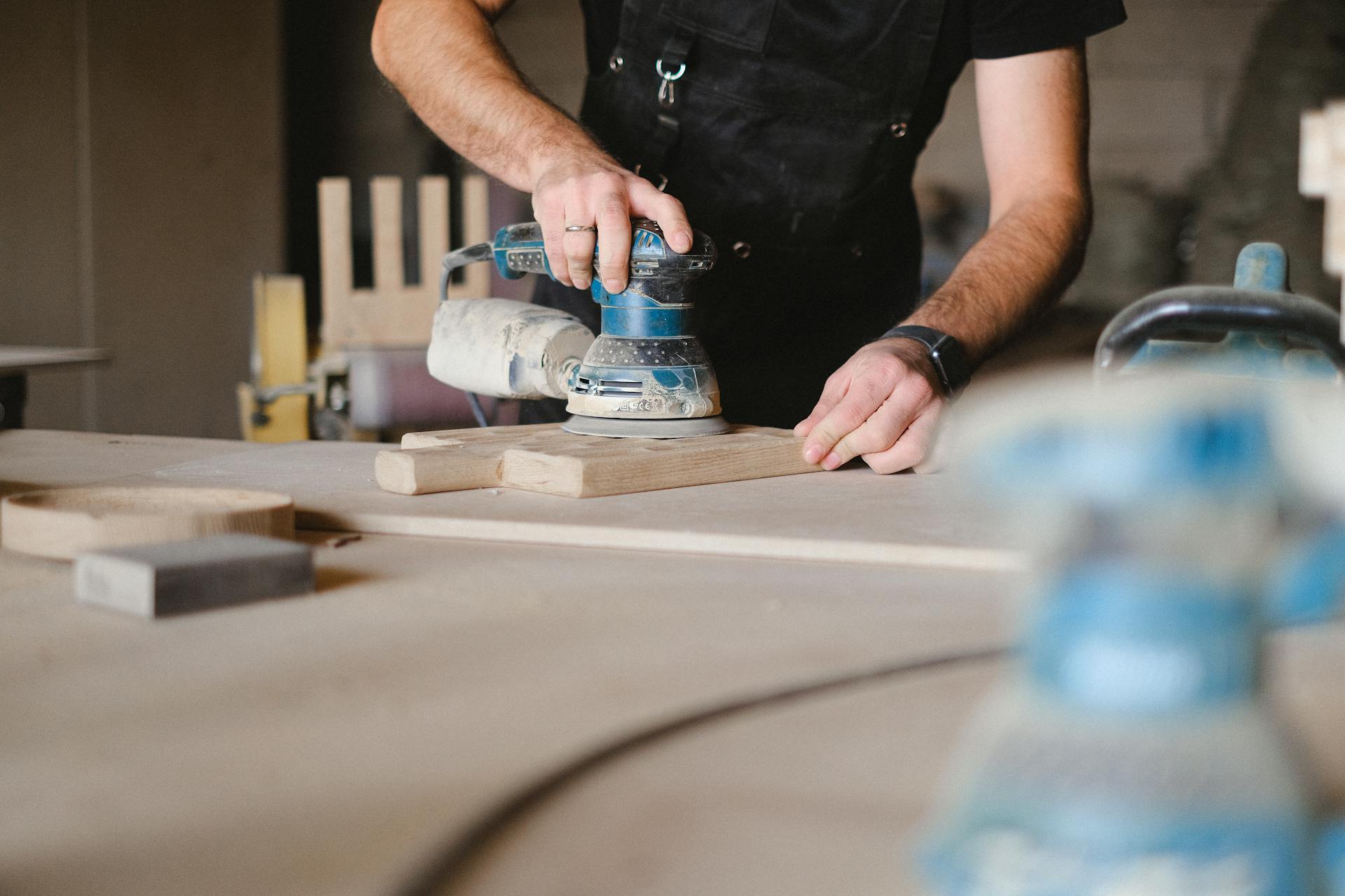 Unrecognizable male woodworker wearing uniform and polishing wooden board with random orbital sander at big table in professional studio