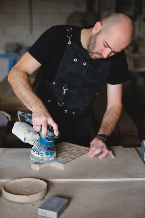 From above of serious bearded male woodworker polishing small wooden board with special instrument while standing at workbench during work in professional joinery