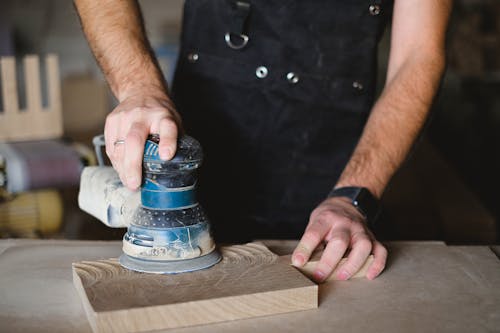 Unrecognizable male carpenter using random orbital sander while working with wooden detail at workbench in professional workshop  on blurred background