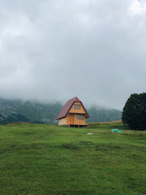 Brown Wooden House on Green Grass Field Near Green Mountains Under White Clouds