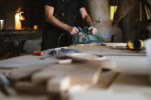 Unrecognizable male artisan using electric belt sander while working at workbench with blurred stack of wooden planks in professional joinery