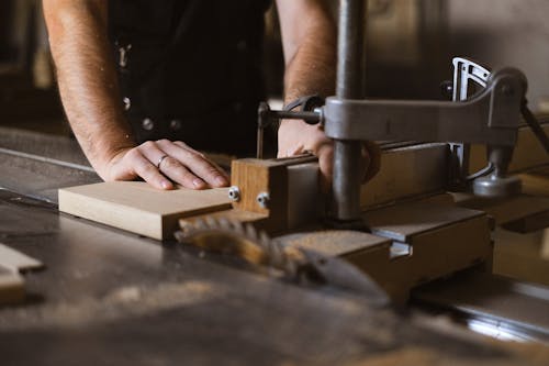 Free Unrecognizable male master cutting piece of wooden plank while working at sawbench in workshop on blurred background with professional equipment Stock Photo