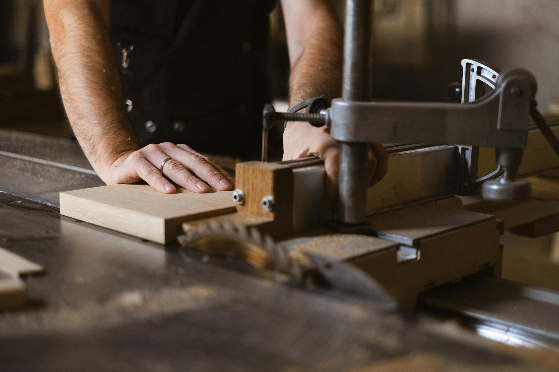 Unrecognizable male master cutting piece of wooden plank while working at sawbench in workshop on blurred background with professional equipment