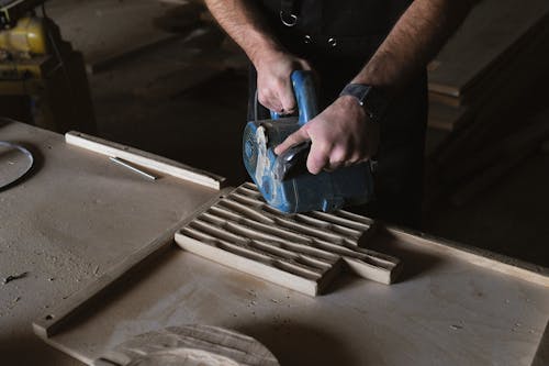 From above of anonymous craftsman using special electric tool to polish wooden board while working at table in professional joinery