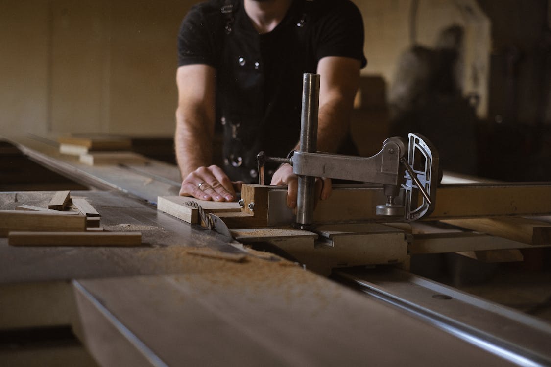 Woodworker in apron standing at sawbench while working with wooden details in professional carpentry with stack of wood