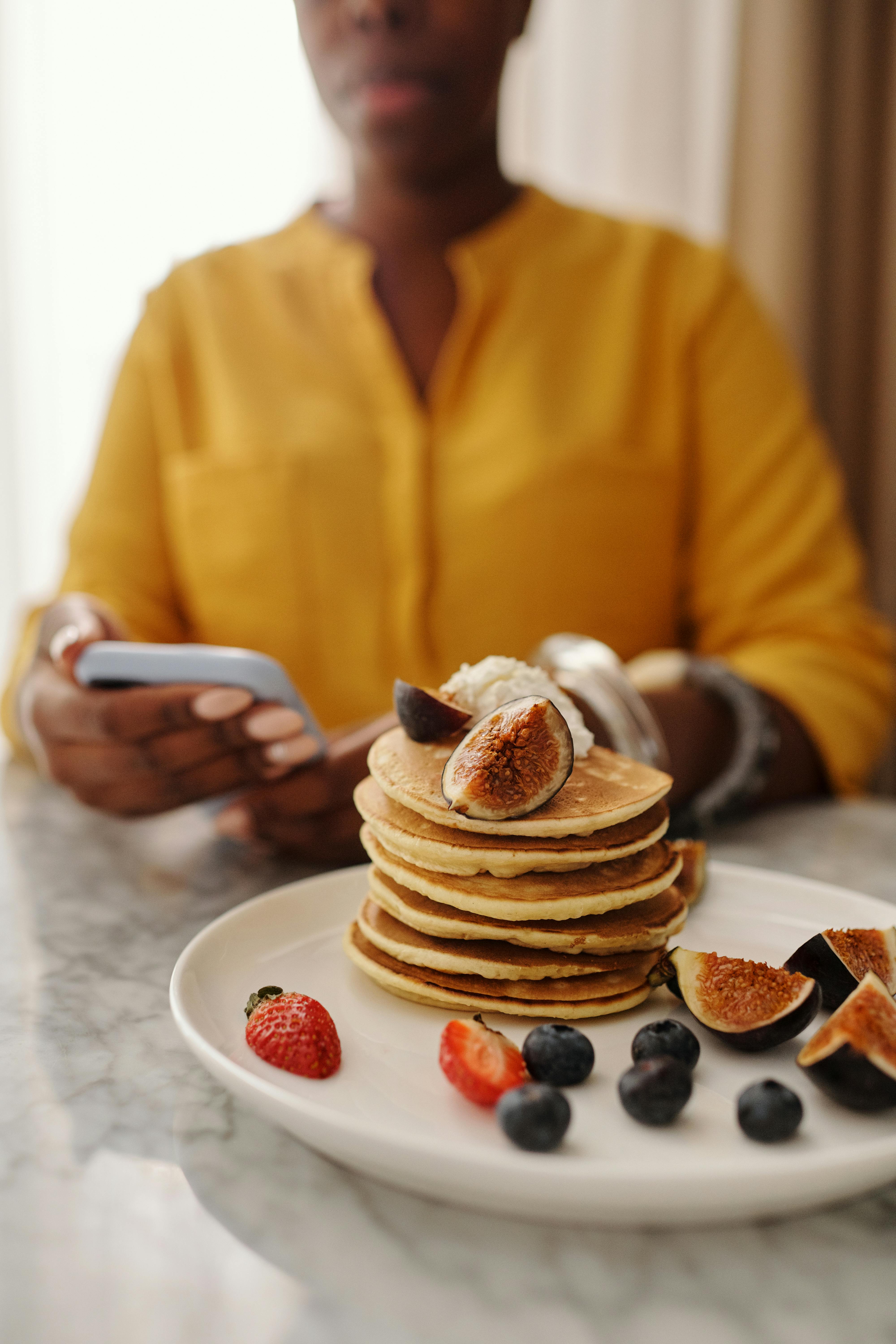Stack of pancakes topped with figs and berries, person in background using phone.