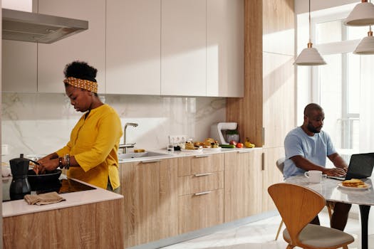 Couple in a modern kitchen, one cooking breakfast and the other using a laptop.