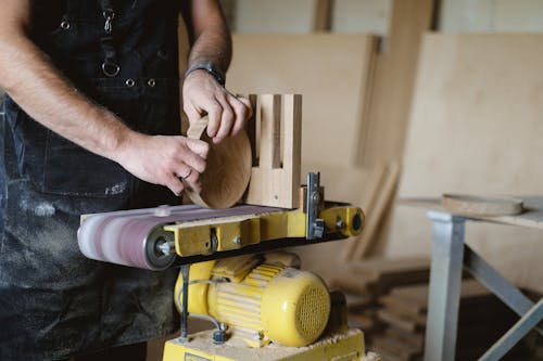 Anonymous male master using belt and disc sander to shape piece of wood  while working in professional carpentry with stack of wood