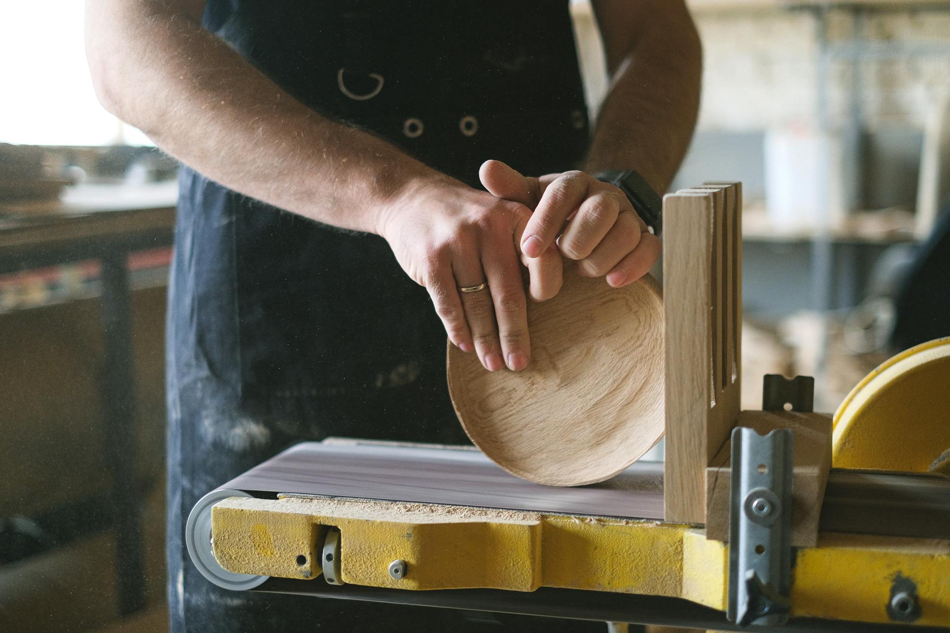 Unrecognizable male woodworker shaping round wooden detail while working at belt and disc sander in professional workshop on blurred background