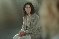 一位戴着眼镜、穿着格子夹克的白人妇女坐在室内，表情沉思