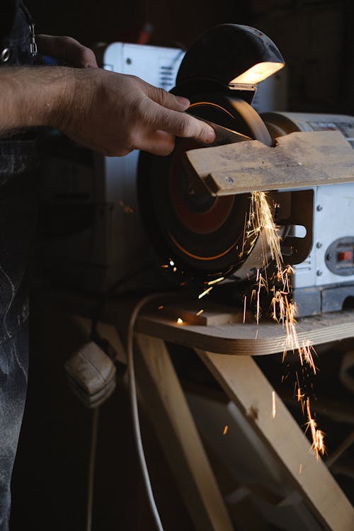 Unrecognizable male master using grinding machine with sparks to sharpen wooden board while working at workbench in professional joinery with instruments