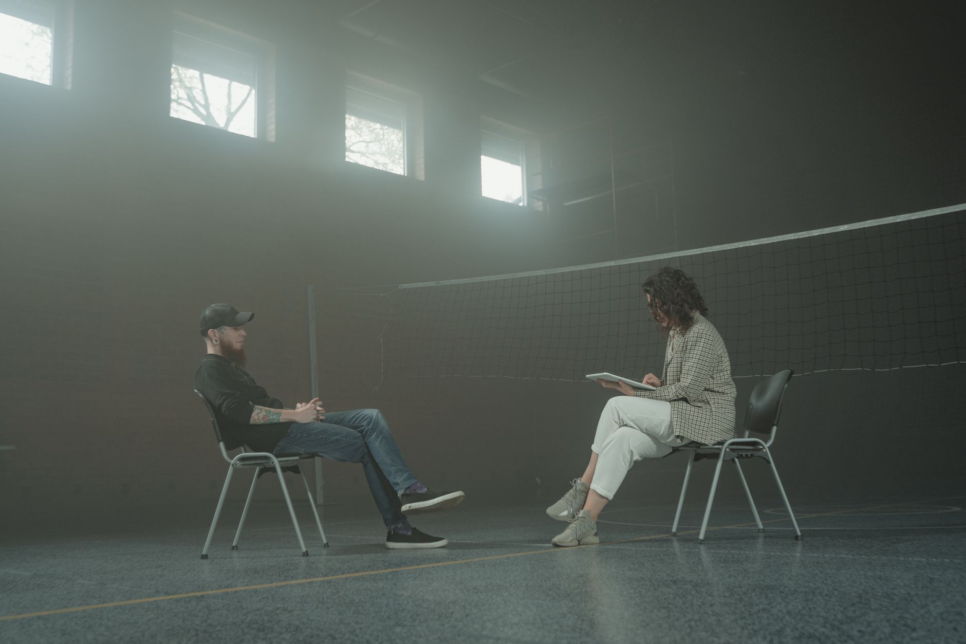 A bearded man and a woman sit in chairs during a counseling session in a dimly lit gymnasium.