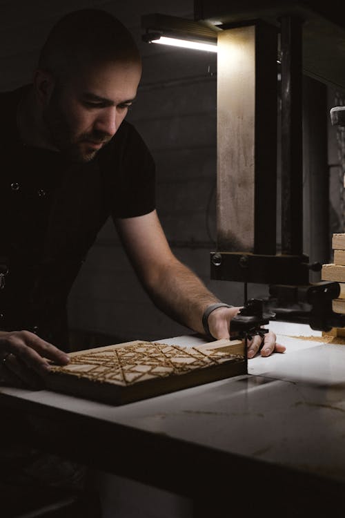 Free Concentrated male joiner using bandsaw machine while creating patterns on wooden board in dark workshop with special instruments and glowing lamp Stock Photo