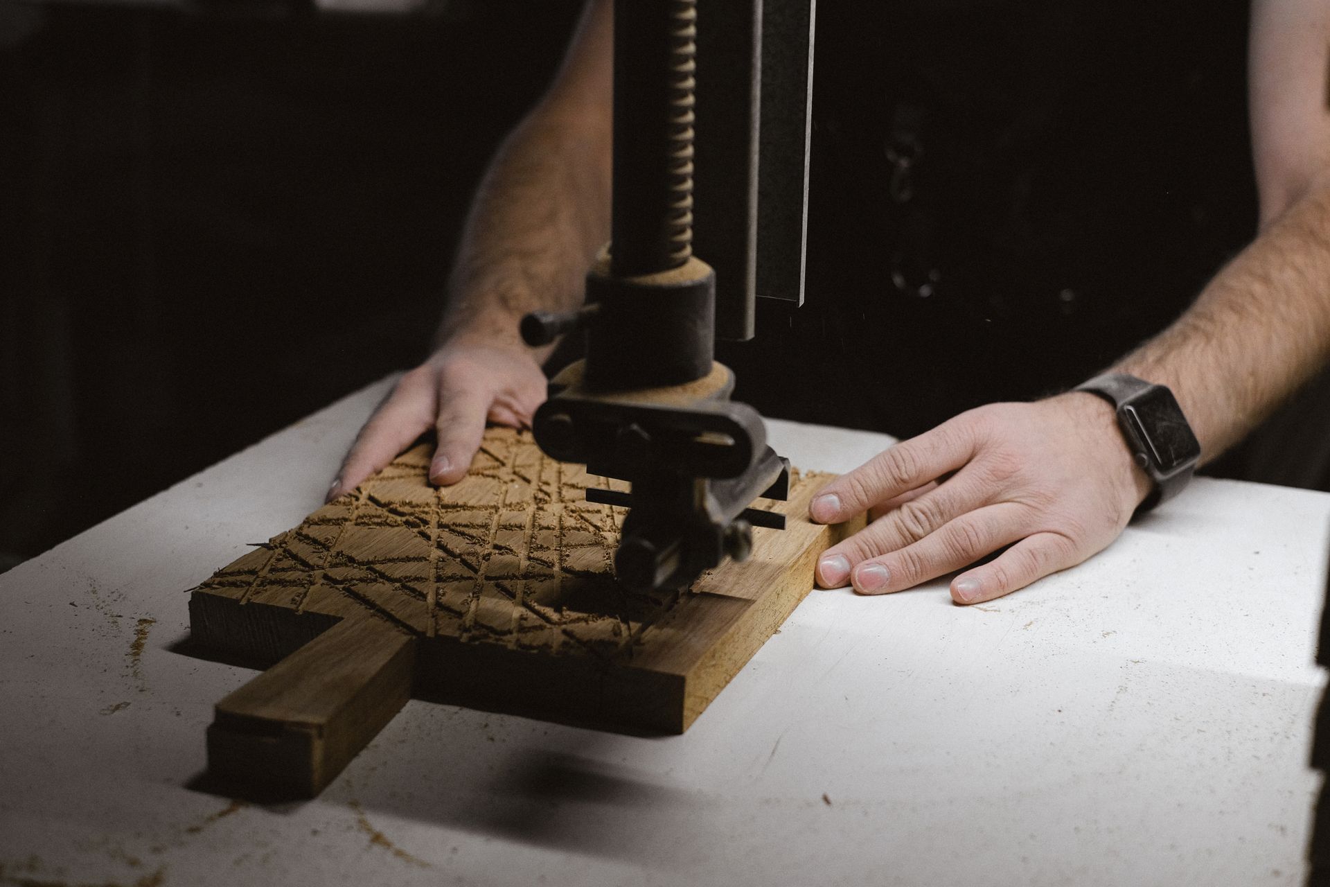 Unrecognizable male carpenter using special metal instrument to carve wooden board while working at workbench on black background in dark workshop