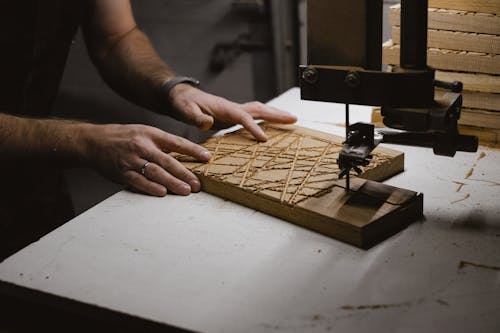 Unrecognizable male master using sharp bandsaw machine while creating  pattern on wooden plank in dark professional carpentry with equipment