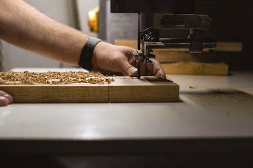 Unrecognizable male carpenter using band saw machine while creating ornament on wooden board at workbench in professional joinery with special equipment