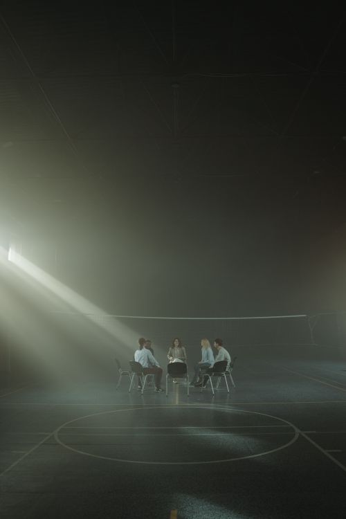 Free A group of adults sitting in a circle discussing under soft lighting in a gymnasium. Stock Photo