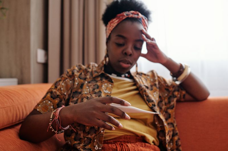 A Black woman in a patterned shirt and headwrap sits on an orange couch at home, one hand pressed to her forehead and her eyes lowered, holding a digital thermometer.