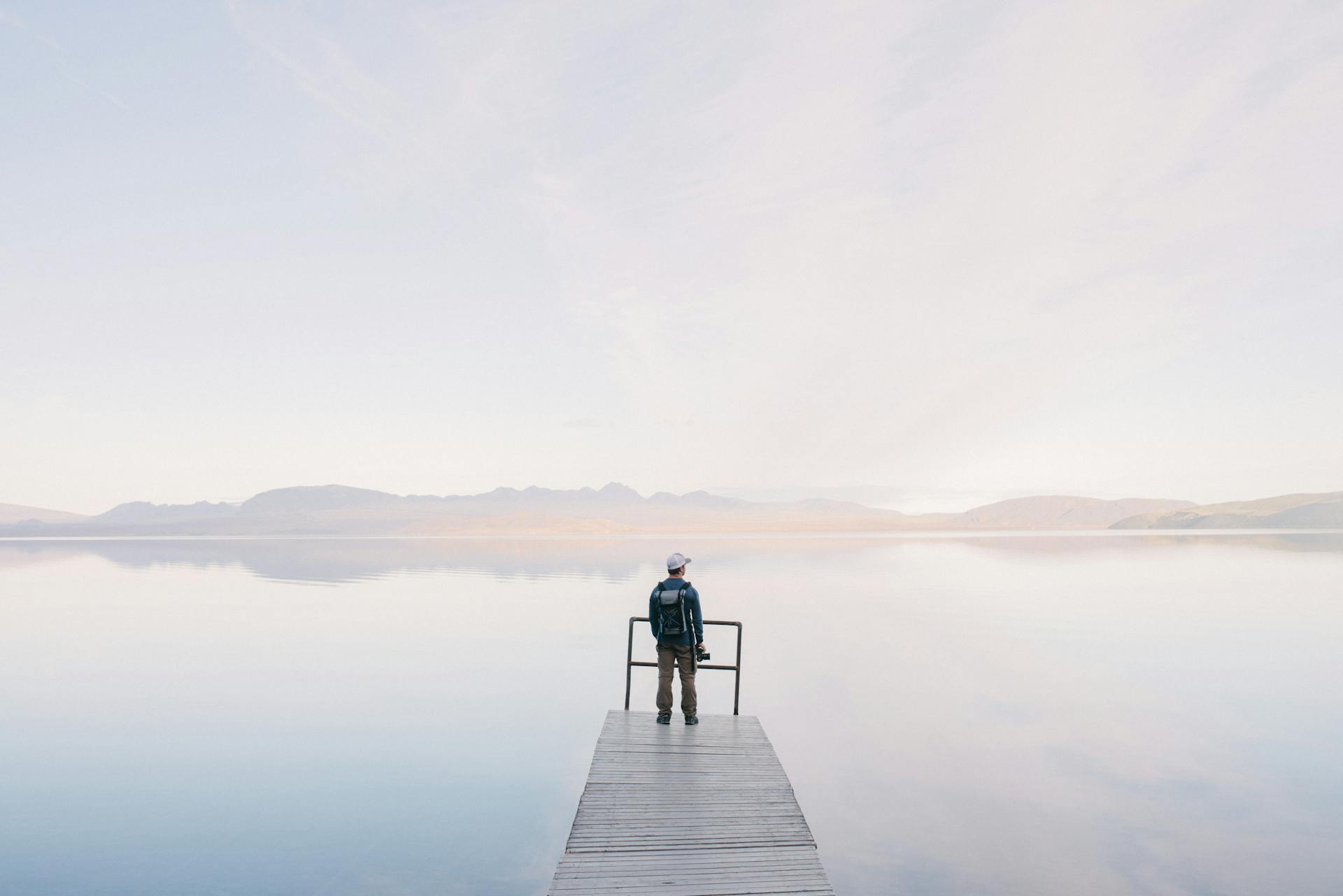 Man Wearing Jacket Standing on Wooden Docks Leading to Body of Water