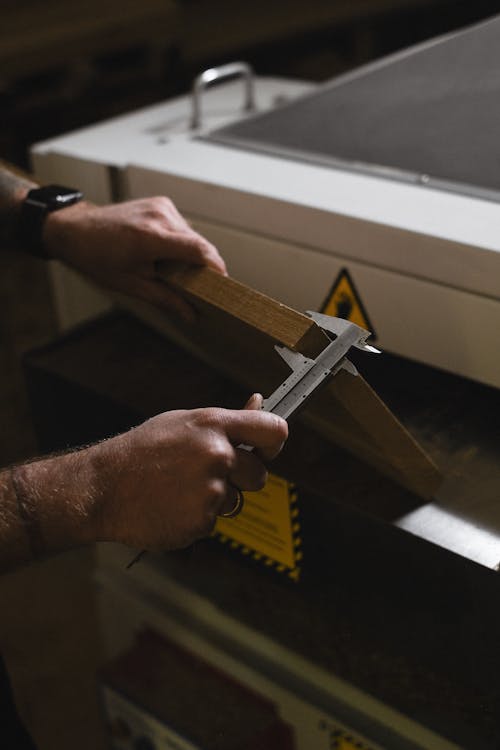 Unrecognizable male artisan using vernier caliper to measure wooden plank while standing near special machine in professional joinery during work
