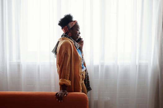 Stylish woman with headband talks on smartphone, standing by a bright window.
