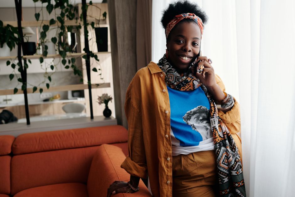 Cheerful woman in a colorful outfit talking on her smartphone, standing next to an orange sofa in a well-lit living room.