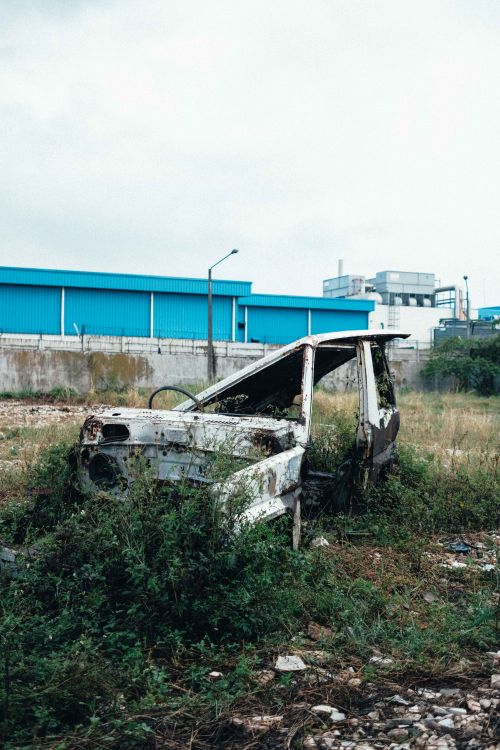 Free Rusty, abandoned car among overgrown weeds, set against industrial buildings in Bandung, Indonesia. Stock Photo