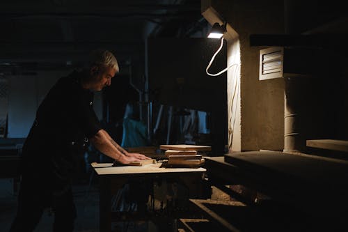 Side view of skilled male master processing wooden board while working at workbench in dark workshop with glowing lamp